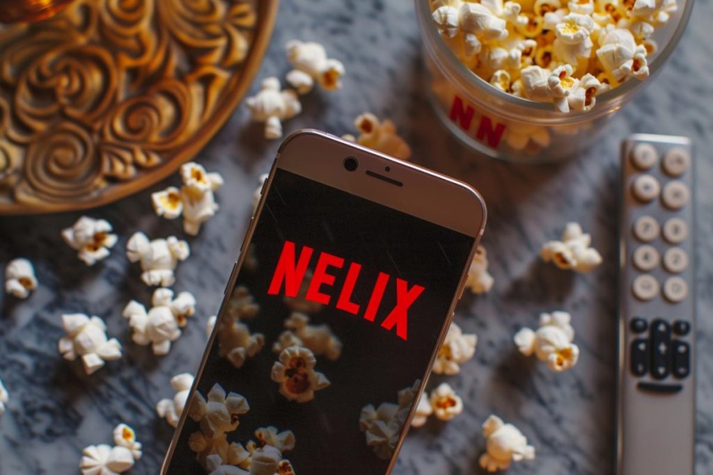 Smartphone affichant « NELIX » à l'écran entouré de pop-corn dispersés, d'un contenant de pop-corn et d'une télécommande de télévision sur une table.