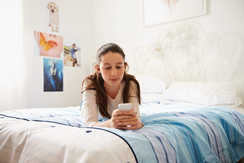 Une fille est allongée sur un lit et utilise un smartphone. La chambre est décorée d'un couvre-lit à motifs et de photos sur le mur.