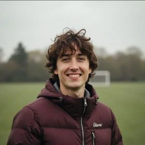 Une personne aux cheveux bruns ondulés, vêtue d'une veste polaire bordeaux, se tient à l'extérieur sur un terrain herbeux avec un but de football et des arbres en arrière-plan.