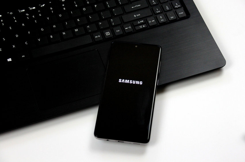 Un smartphone Samsung avec le logo sur son écran repose sur un clavier d'ordinateur portable noir, les deux appareils étant placés sur une surface blanche.