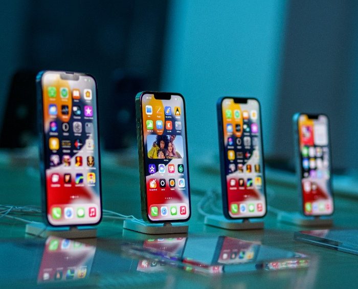 Quatre smartphones sont présentés à la verticale sur un comptoir, chacun affichant un écran d'accueil avec diverses icônes d'applications sous un éclairage intense.