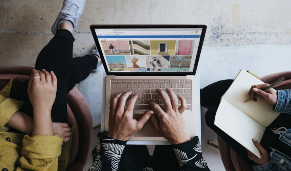 Trois personnes sont assises autour d'une table ; l'une tape sur un ordinateur portable affichant des vignettes d'images, l'autre tient un carnet et un stylo, et la troisième pose ses mains sur ses genoux.