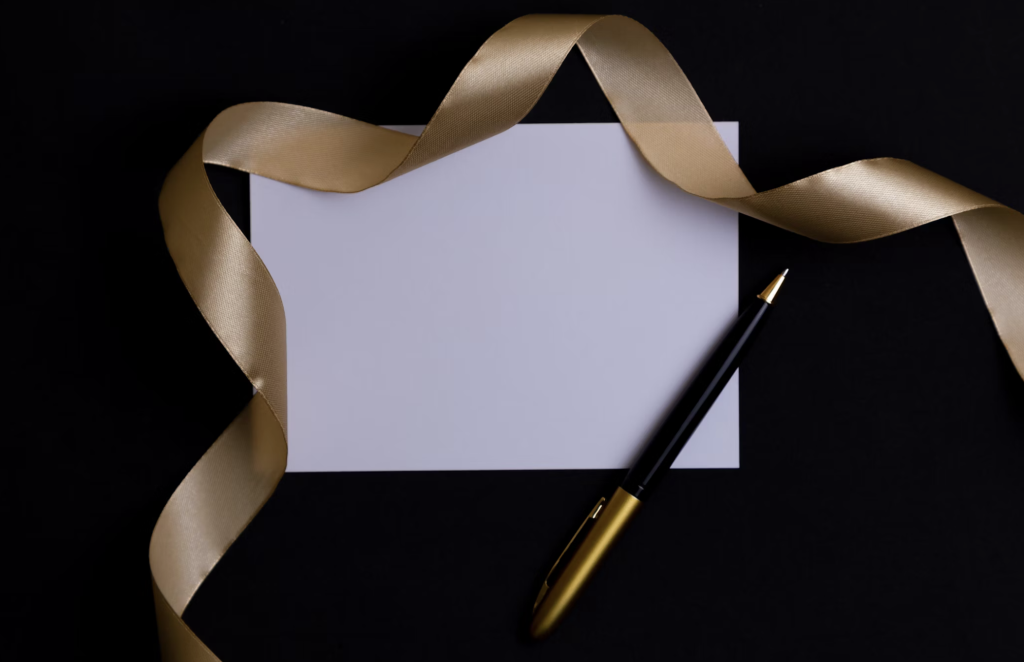Une carte blanche vierge avec un ruban doré et un stylo noir et or sur fond noir.