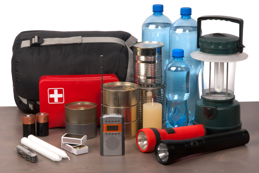 Un kit d'urgence comprenant une trousse de premiers secours, des conserves, des bouteilles d'eau, des lampes de poche, des piles, une bougie, des allumettes, une lanterne, une radio et un sac de couchage sur une table.