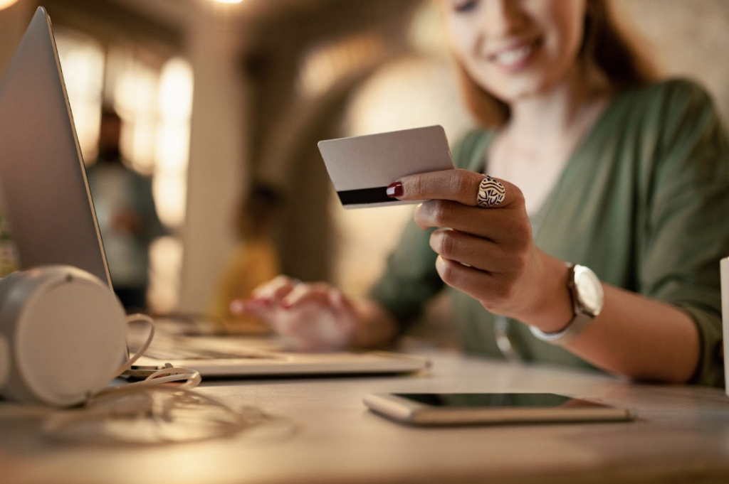 Une femme est assise à une table, utilisant un ordinateur portable et tenant une carte de crédit, avec un smartphone et des écouteurs à côté d'elle.