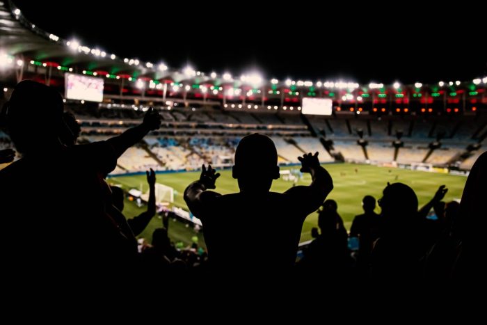 Des supporters silhouettés applaudissent dans les tribunes d'un stade de football illuminé lors d'un match de nuit.