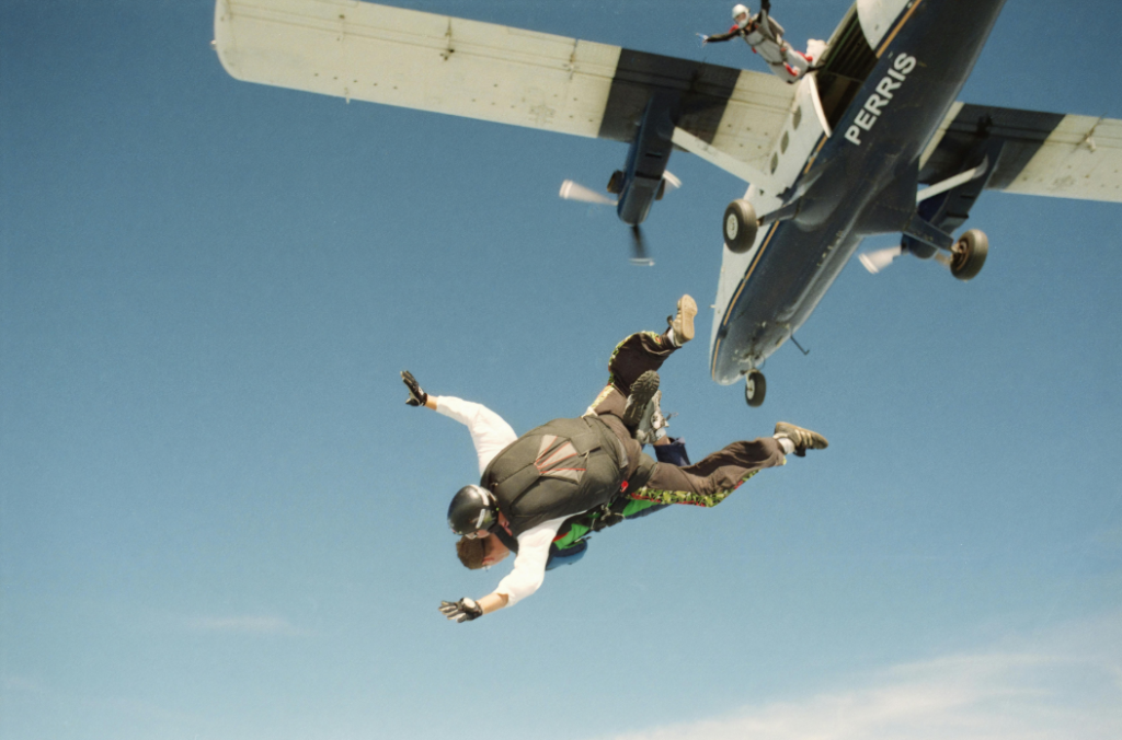 Deux personnes effectuant un saut en parachute en tandem viennent de sortir d'un avion en plein vol sous un ciel bleu clair, un autre parachutiste étant aperçu près de l'avion.