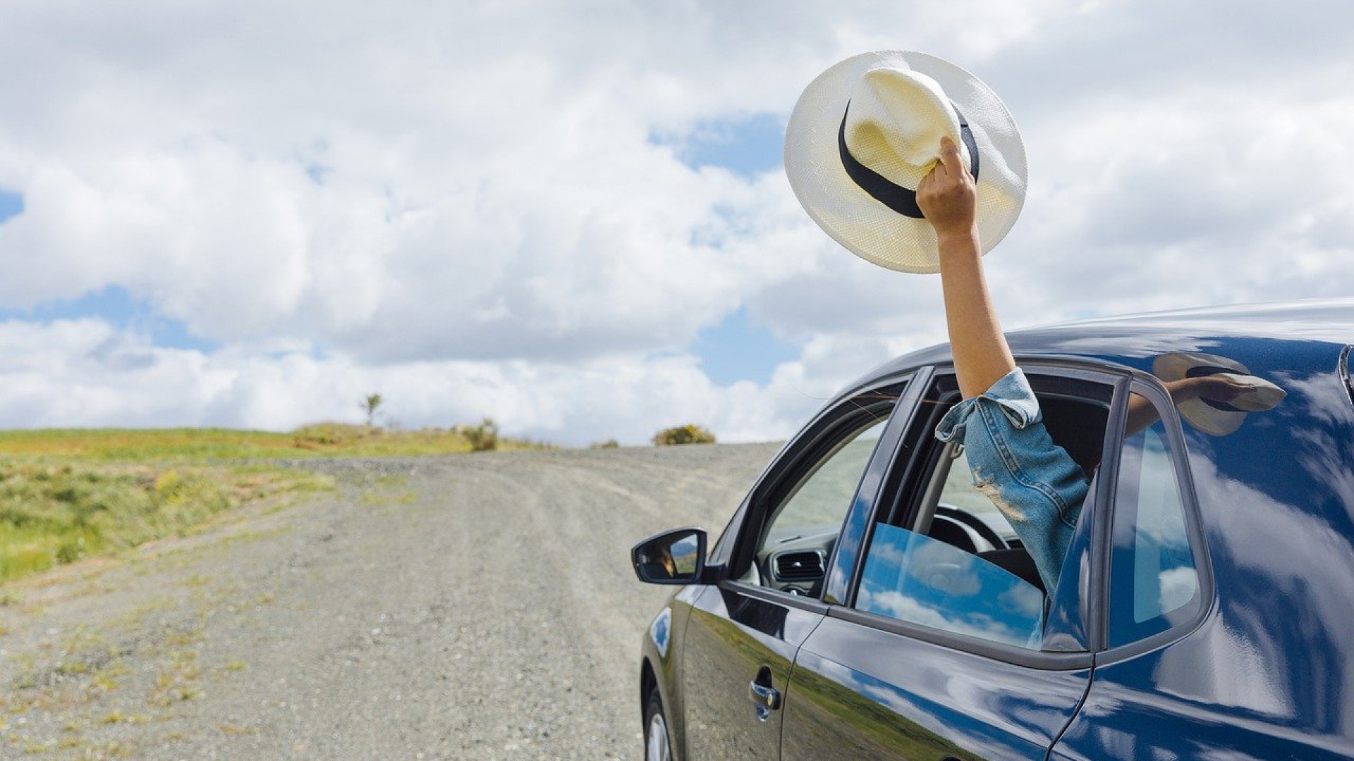 Une personne dans une voiture tient un chapeau blanc par la fenêtre alors qu’elle roule sur une route de gravier sous un ciel nuageux.