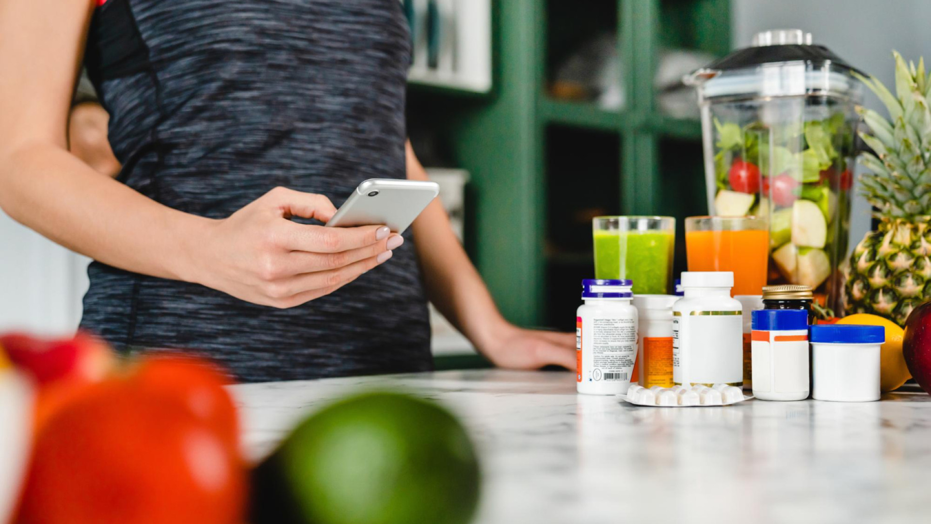 Une personne tenant un smartphone se tient près d&rsquo;un comptoir où sont disposés des fruits, des compléments alimentaires, des comprimés et des smoothies, avec en arrière-plan un mixeur rempli de fruits coupés.