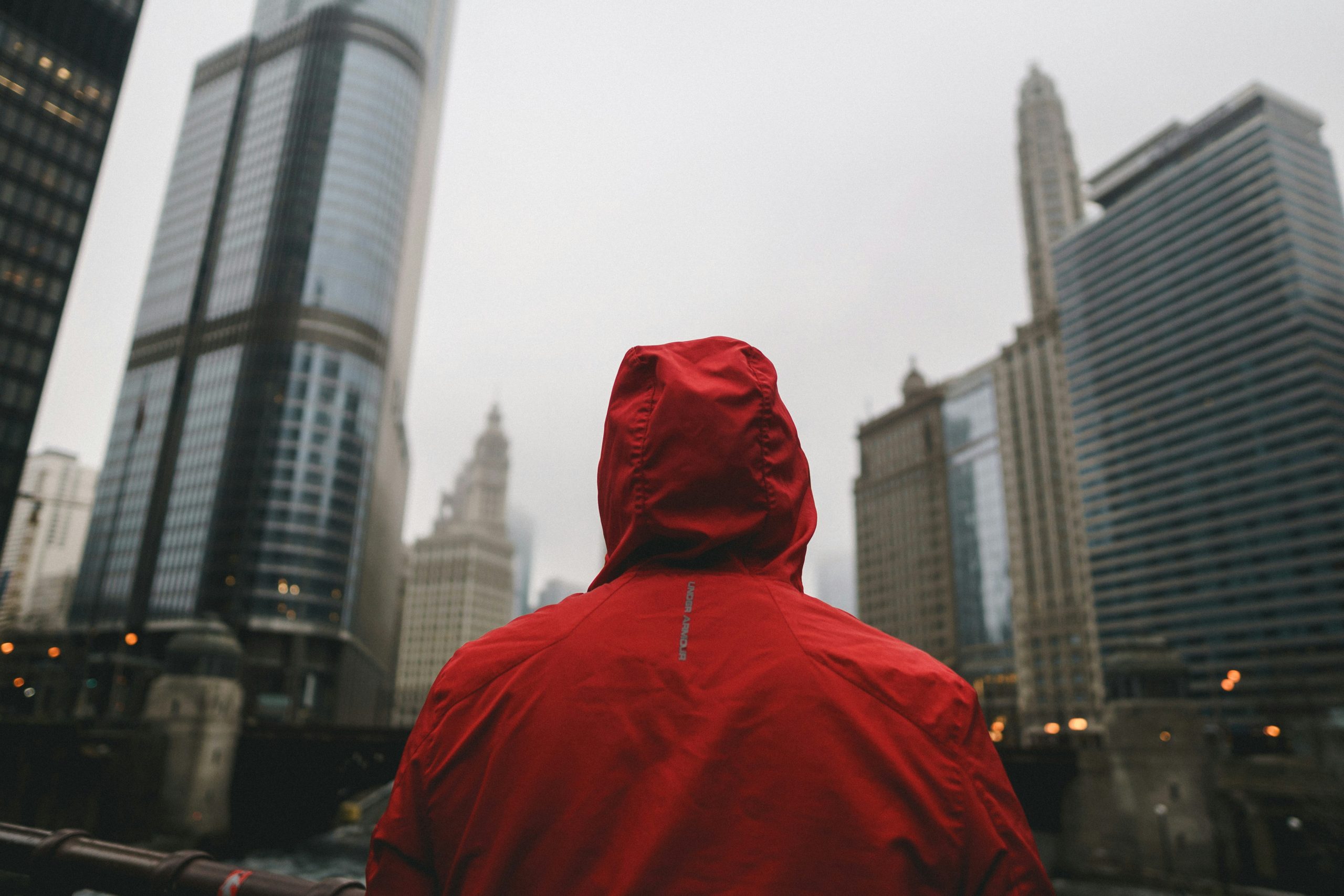 Une personne vêtue d&rsquo;une veste à capuche rouge se tient face à de grands immeubles dans un paysage urbain, par une journée nuageuse.